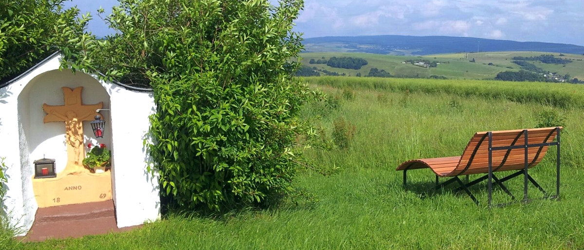 Blick über Franzenheim – weiße Wegkapelle mit Kreuz und Holzbank auf grüner Wiese